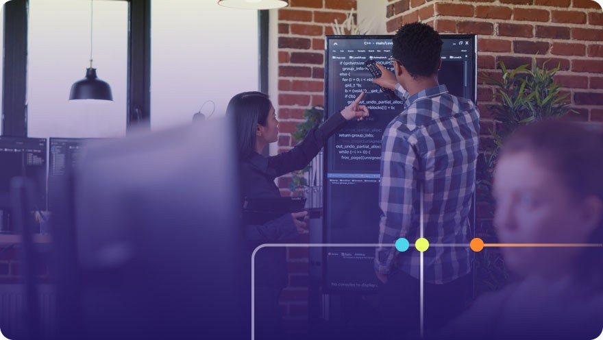 A woman works at a computer in the foreground. In the background, two colleagues are pointing to writing on a chalkboard. A purple overlay with colorful circles.